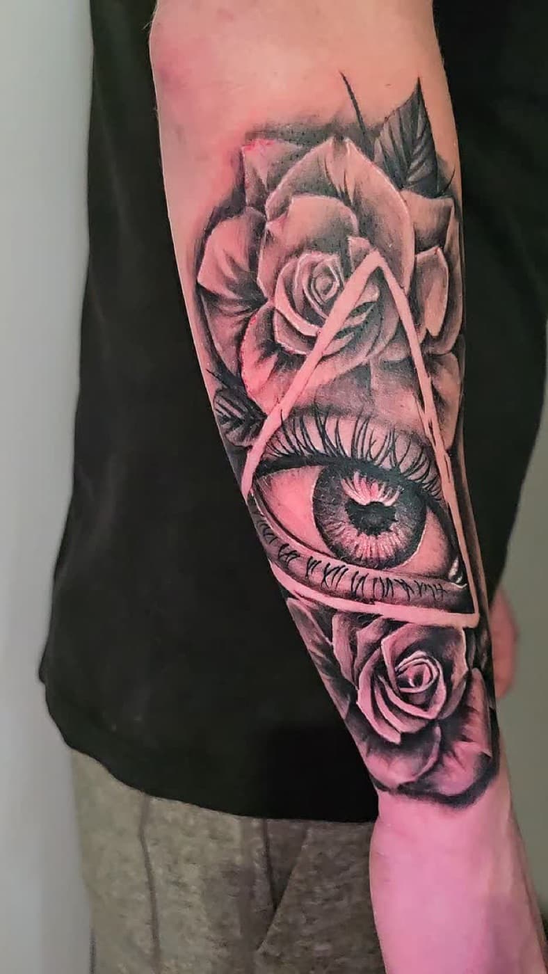 Eye inside a triangle, framed by roses and Roman numerals, black and grey on a forearm
