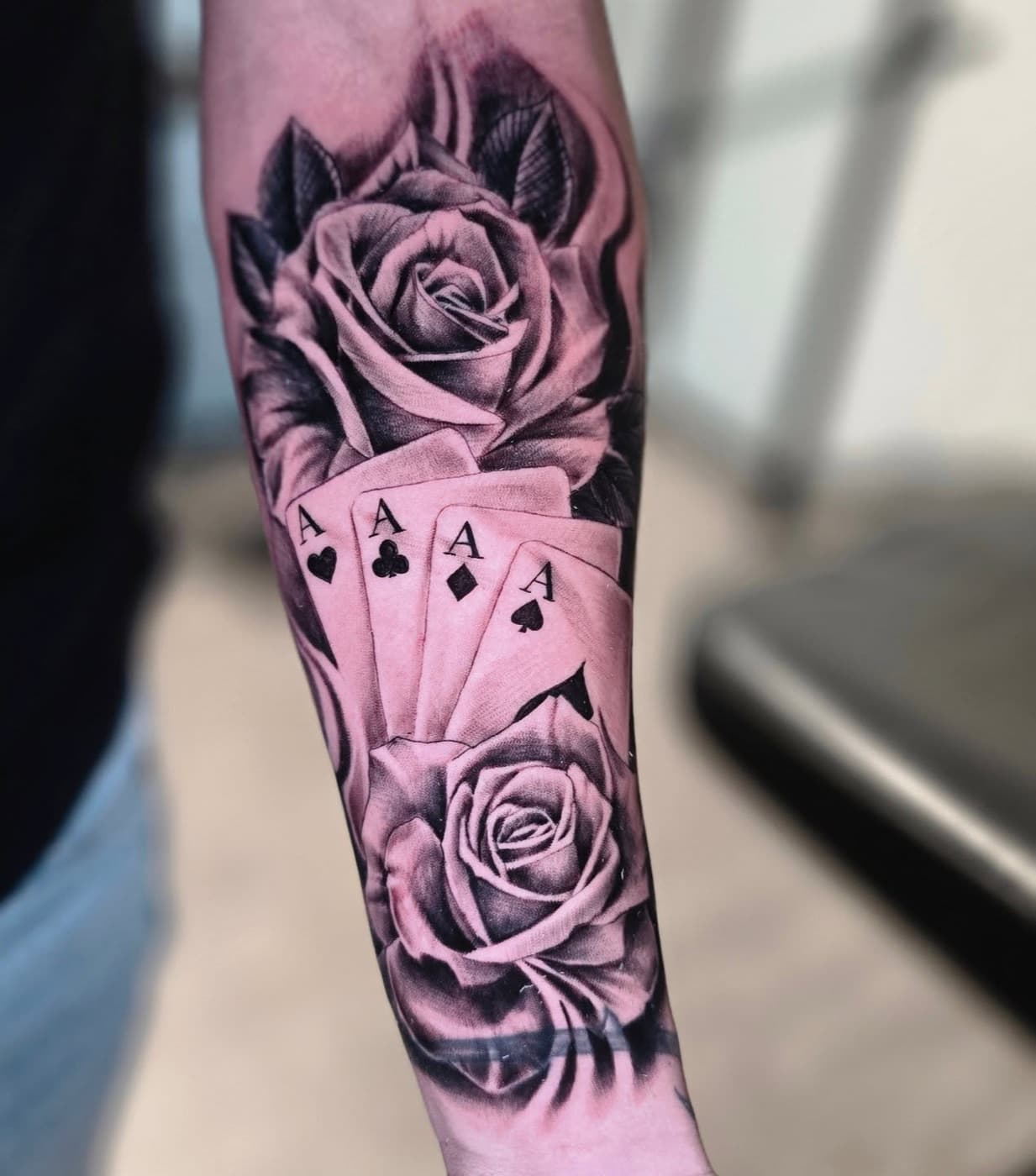 Two roses wrapped around four playing-card aces, black and grey on a forearm