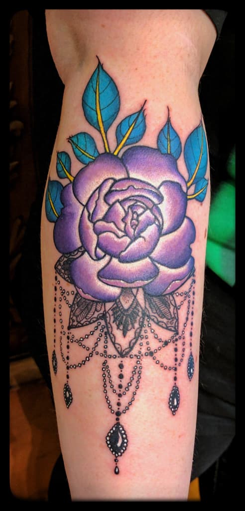 Purple rose with teal leaves above a black lace motif and hanging jewelled chains, colour on the forearm