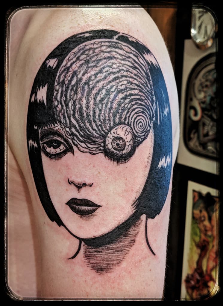 Woman with a black bob and spiral-patterned hair covering one eye that holds an eyeball, black and grey on the upper arm