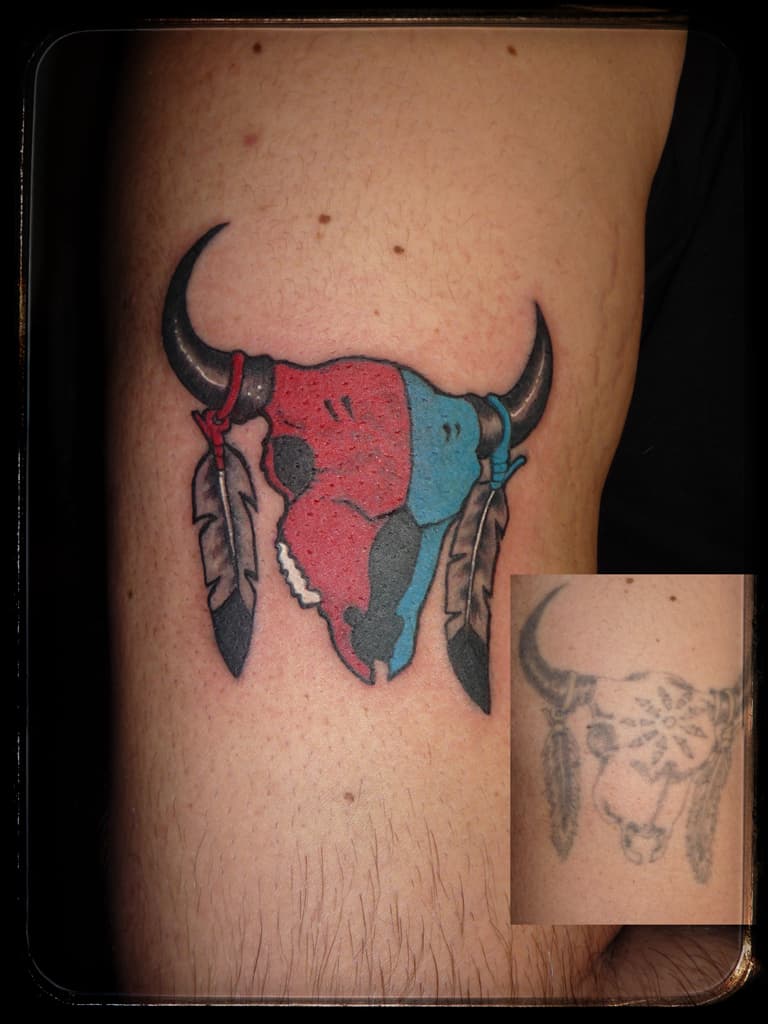Cover-up of a faded bull skull: red and blue colour skull with horns and hanging feathers, the old tattoo inset