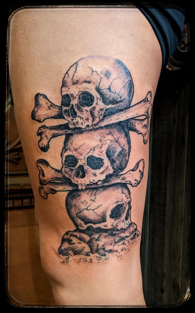 Three cracked skulls stacked with crossed bones on a rock, black and grey on the thigh