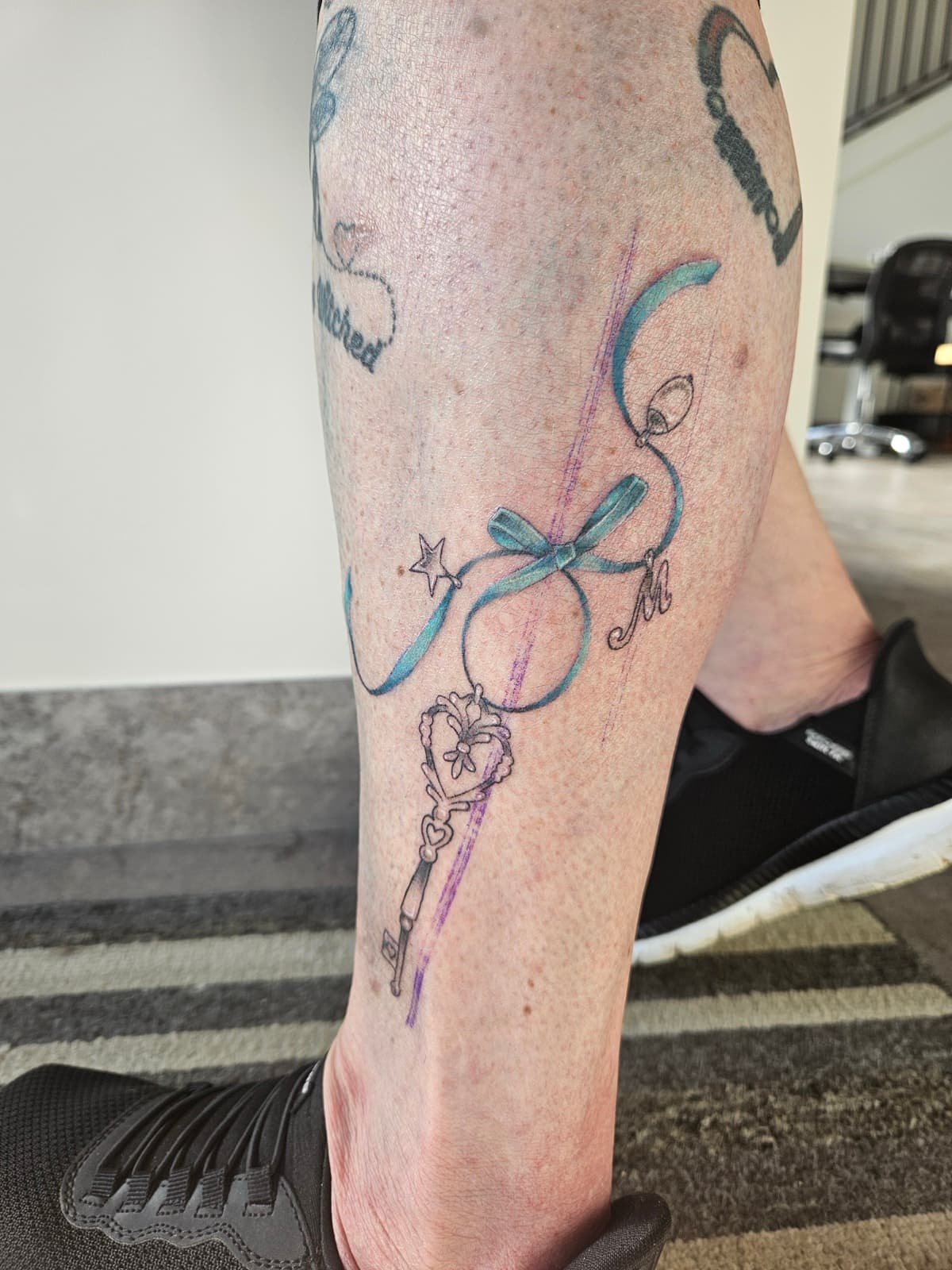 Skeleton key hanging from a teal ribbon bow with a star and an M charm, fine line on the calf