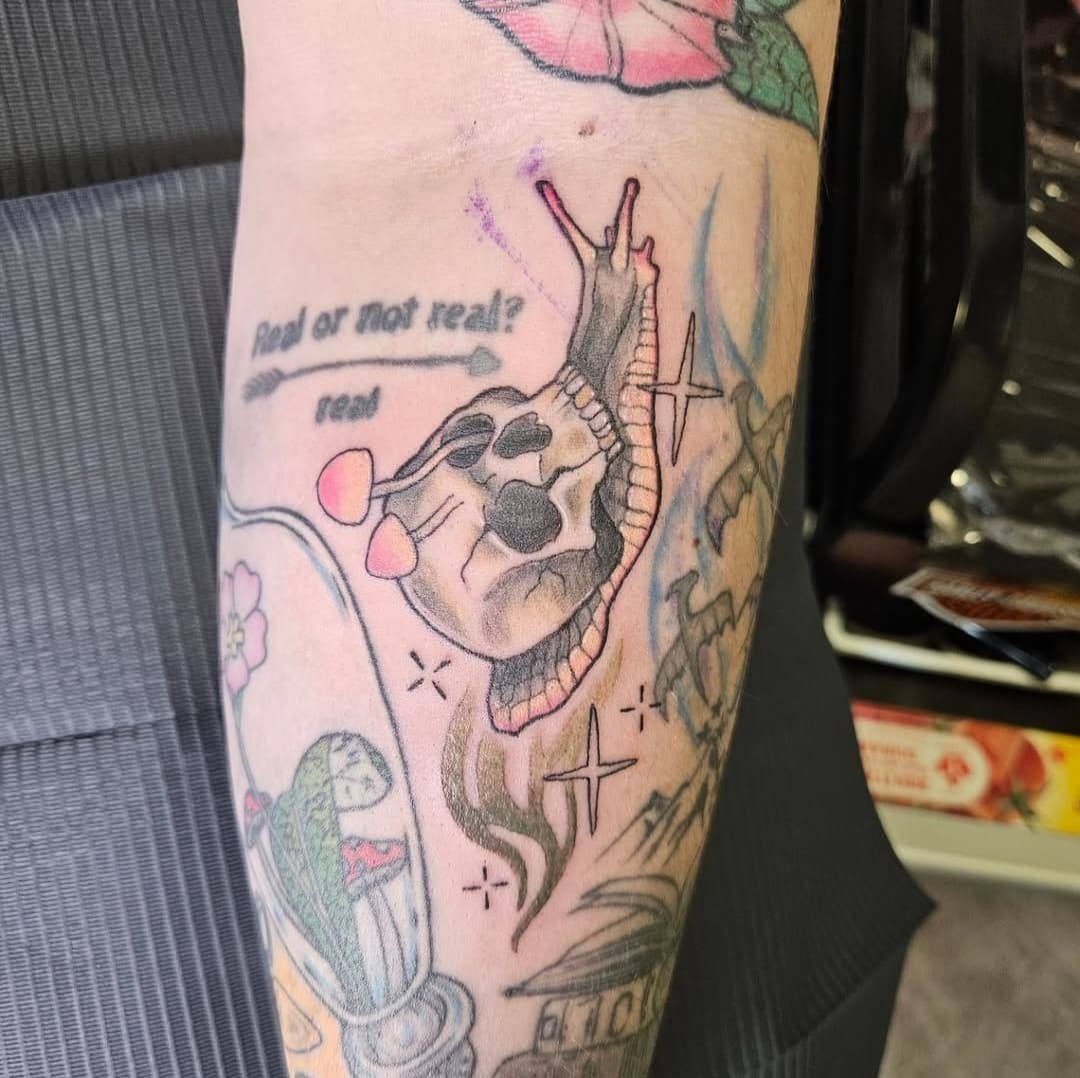Snail carrying a skull as its shell, with pink mushrooms and sparkles, colour on the forearm