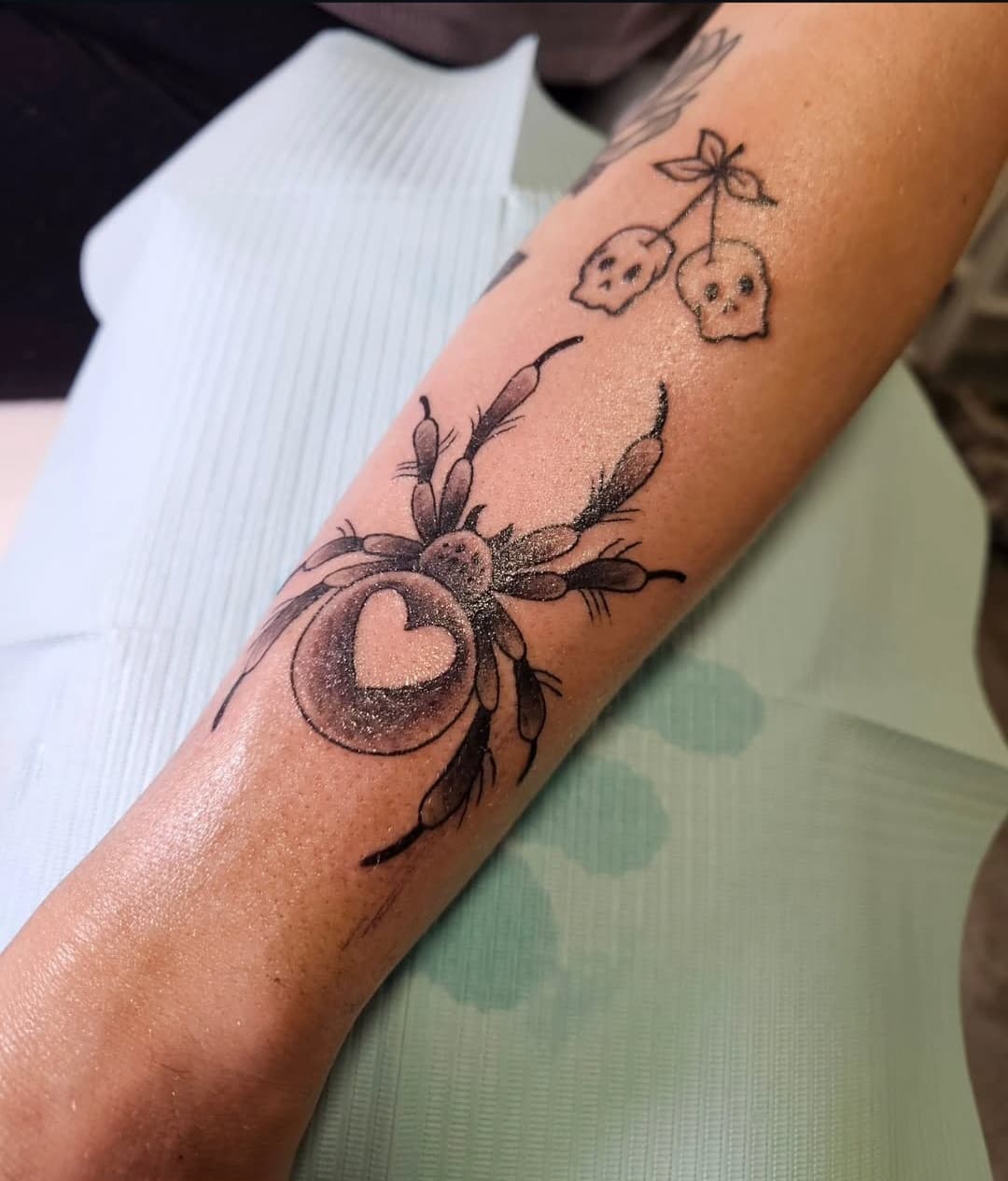 Spider with a heart on its back below two skull cherries, black dotwork on the forearm