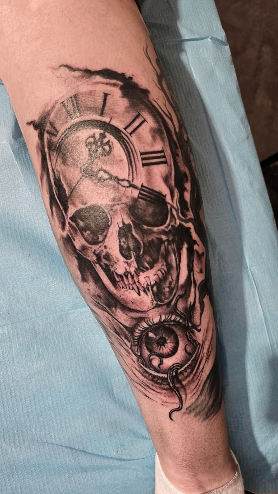 Skull under a cracked pocket watch with Roman numerals and a realistic eye below, black and grey on the forearm